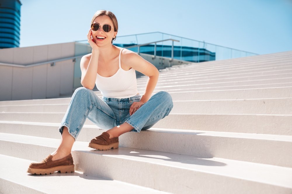 A young woman in sunglasses sits on stairs in a city, wearing jeans, loafers and a white tank top, perfect for summer heat.