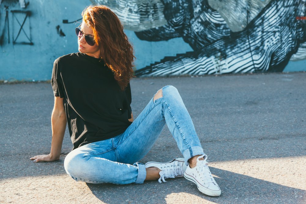 A woman poses on the street with a mural behind her: she wears distressed jeans, white converse, sunglasses and a black t-shirt for an effortlessly stylish and comfortable outfit.