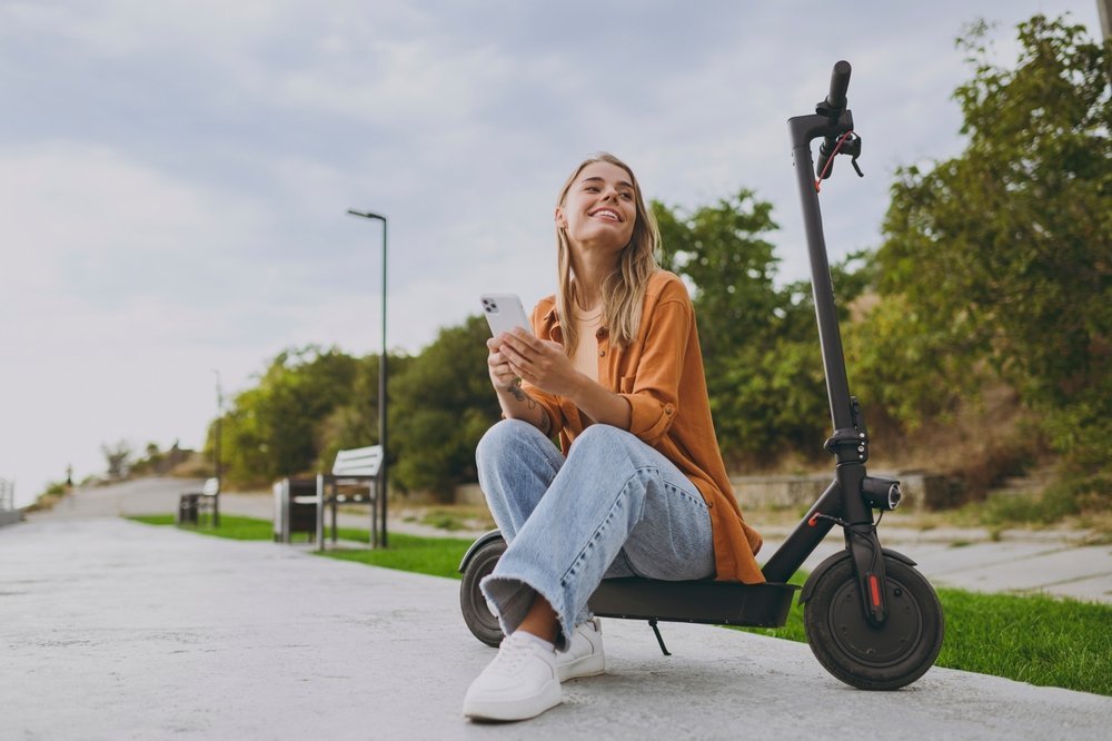 A young blonde girl sits on the edge of a motorized scooter on her phone: she is comfortable in her casual orange t-shirt, linen button up, wide-leg jeans and white sneakers.