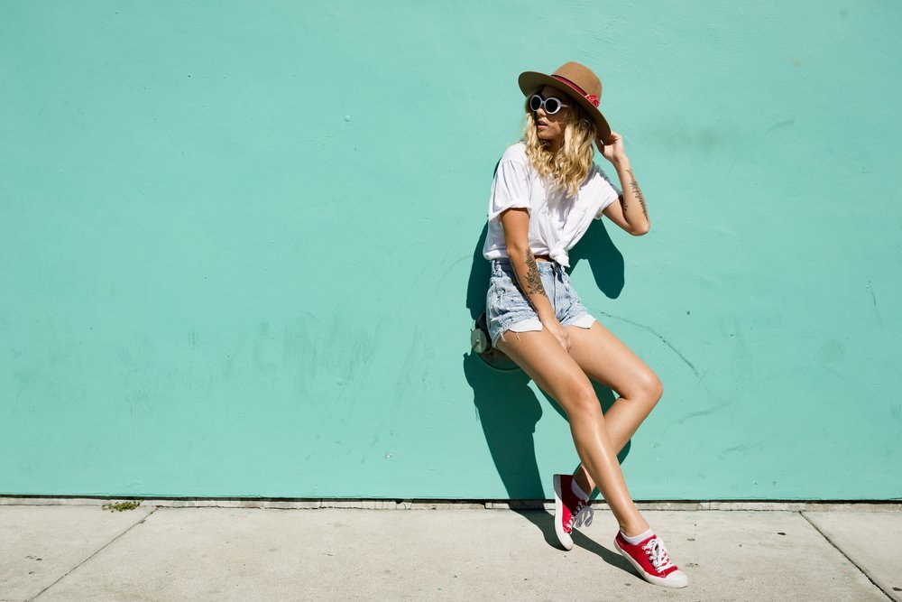 A woman poses against a sea foam green wall in cut off denim shorts, red sneakers, a straw hat, and a white shirt knotted at her belly.