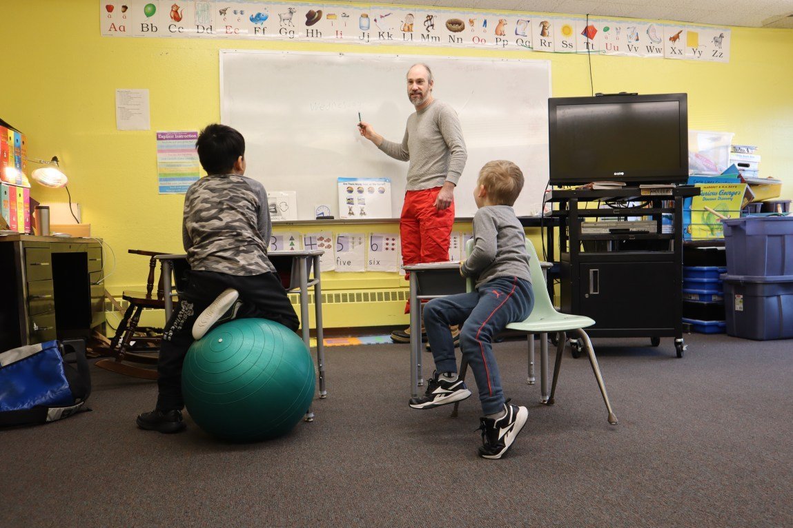 A male teacher stands in front of a white board holding a marker in his hand. Two students sit at school desks in front of him; one sits on a green yoga ball. The classroom has an alphabet poster, a monitor on a rolling cart and various boxes.