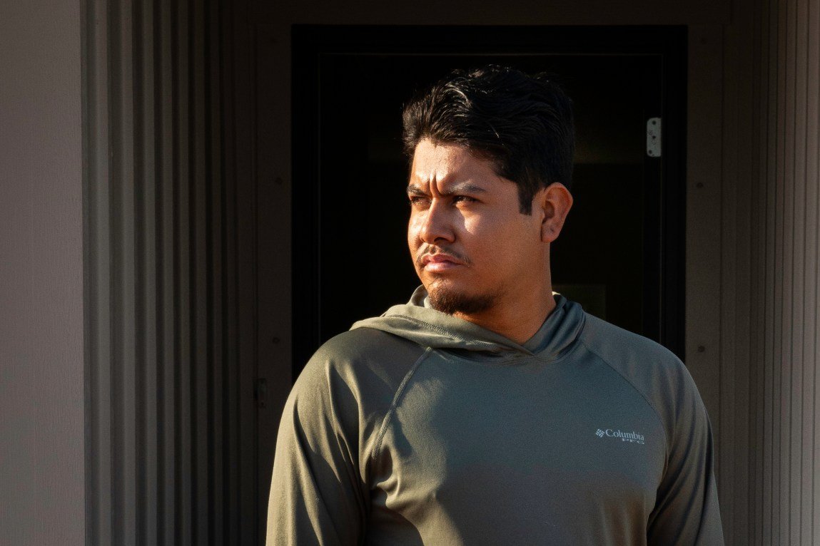 A man in a green hooded shirt looks off into the distance with a somber expression, his face illuminated by the sun as he stands in front of a dark doorway.