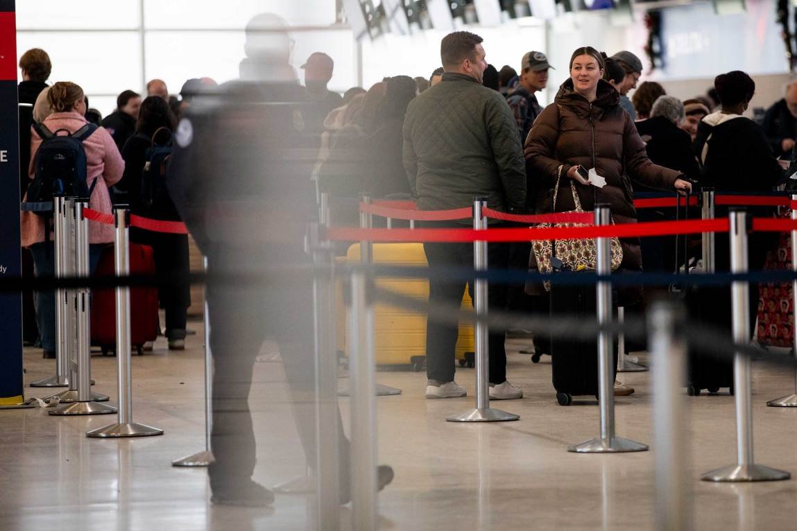 A busy airport terminal where travelers wait in a long, winding line, with the blurred silhouette of a passenger passing by in the foreground.