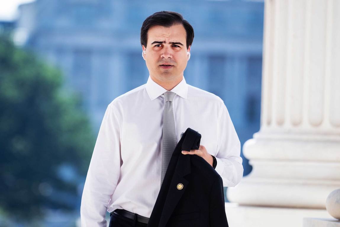 A man wearing a white dress shirt, a gray tie and wireless earbuds holds a dark suit jacket over his arm, next to a large marble column.