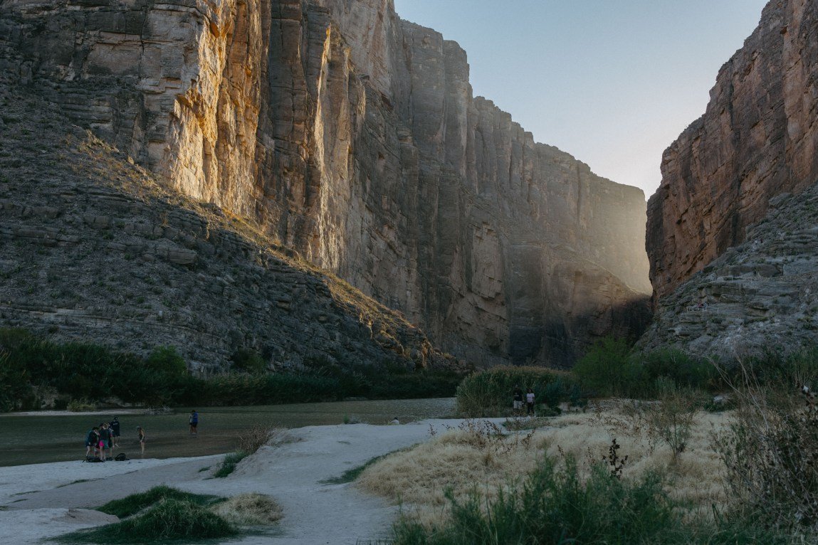 A view of Santa Elena Canyon in Big Bend National Park showing towering rock cliffs framing a river with people on a sandy bank in the foreground.