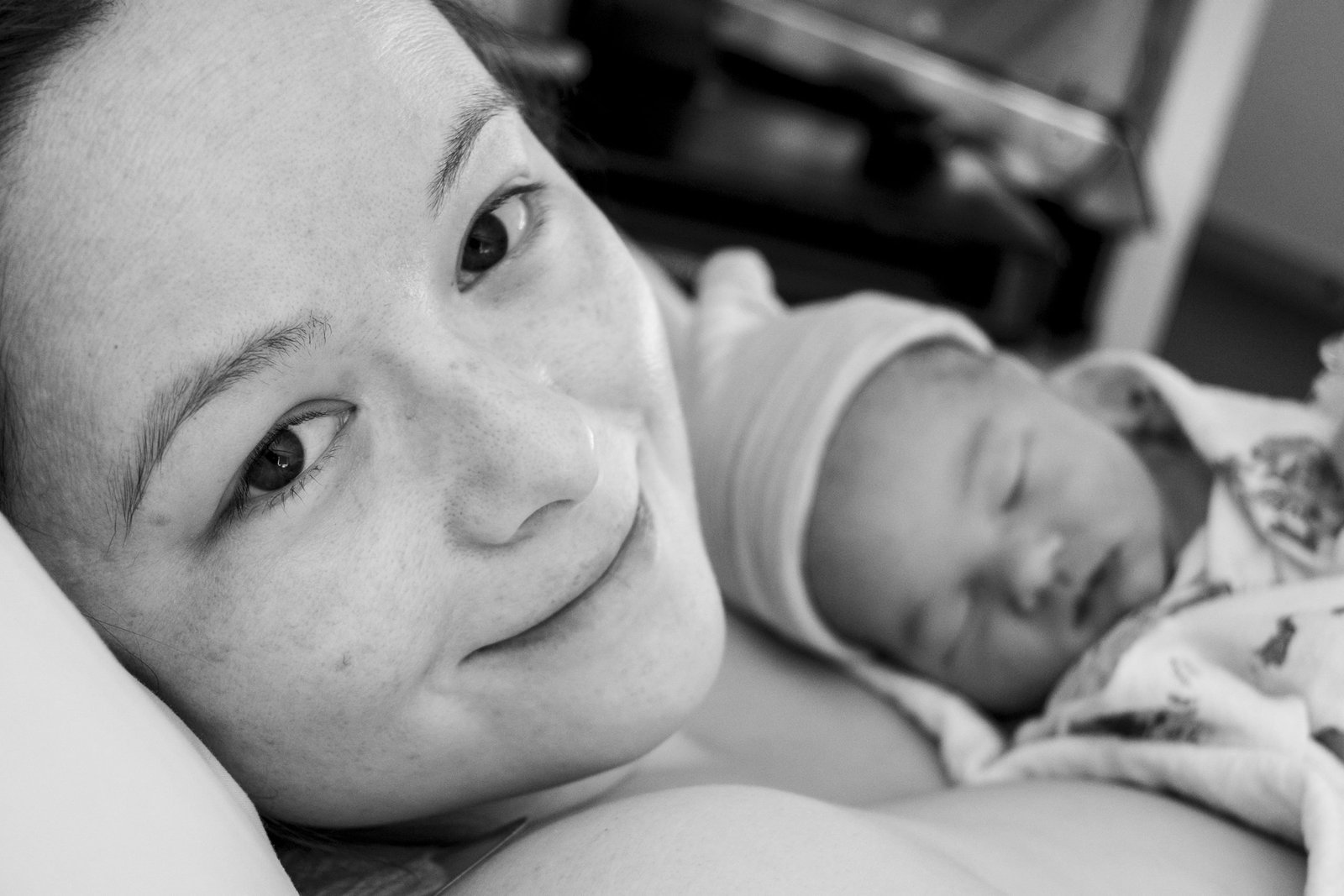 A woman stares directly into the camera smiling and lying on a pillow. An out of focus baby wearing a hat and swaddled in a blanket lies on her chest.