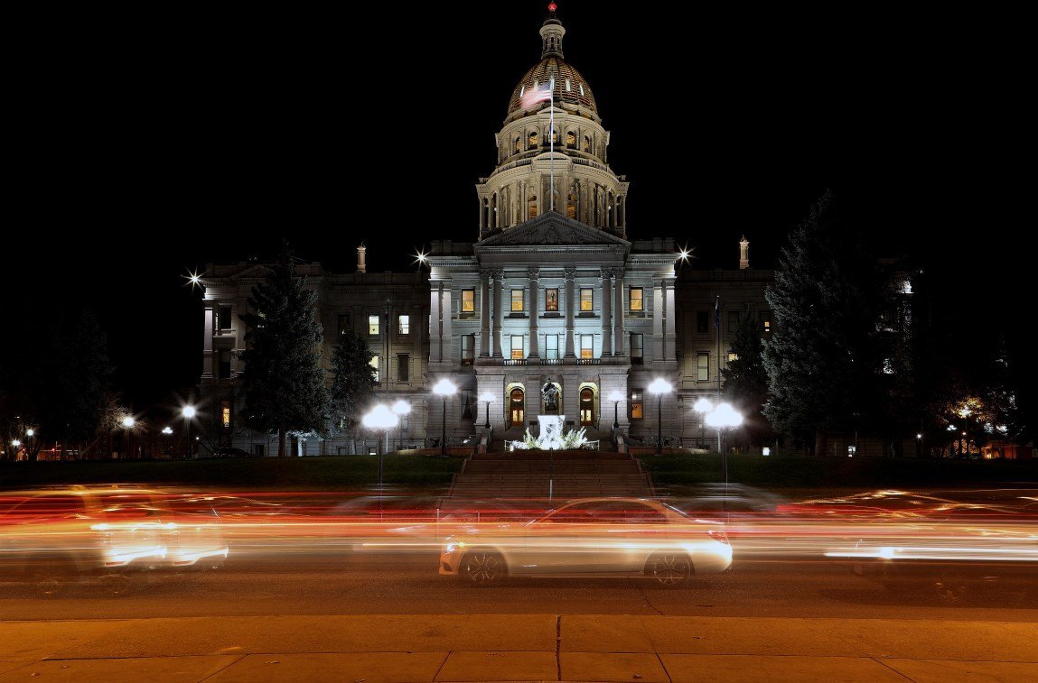 The Colorado State Capitol lit up at night. In the foreground, cars speed by as their headlights and taillights form long streaks of color.