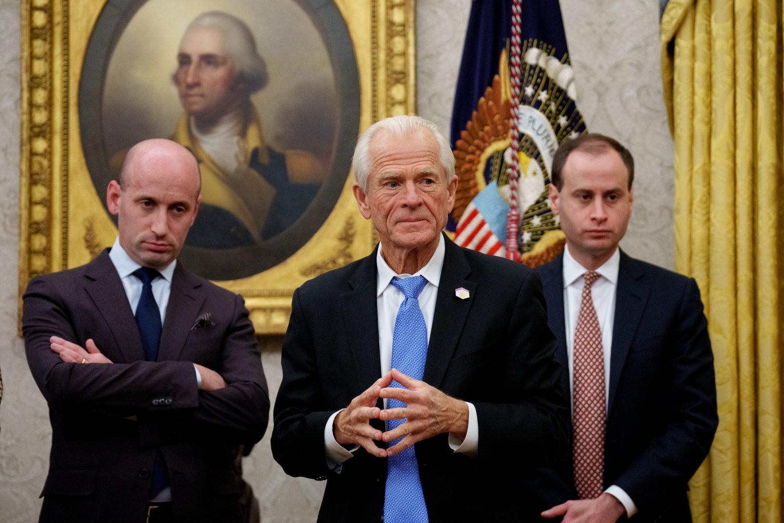 Three men in suits stand in the Oval Office. The man in the center has his fingertips touching. Behind them is a painting of George Washington in an ornate gold frame.