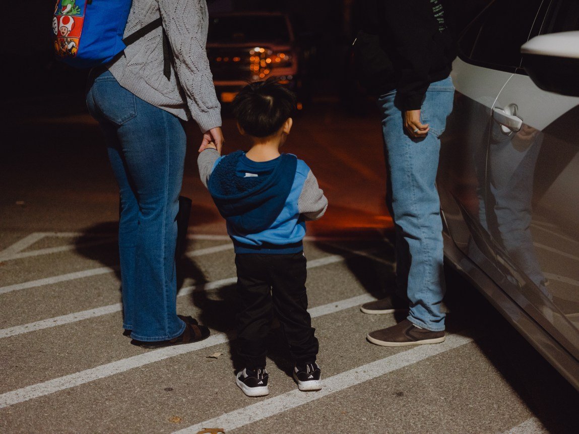 A young boy in a blue and gray hoodie stands in a parking lot at night, holding the hand of an adult in jeans and a gray sweater on the left. Another person in jeans stands on the right next to a white car.