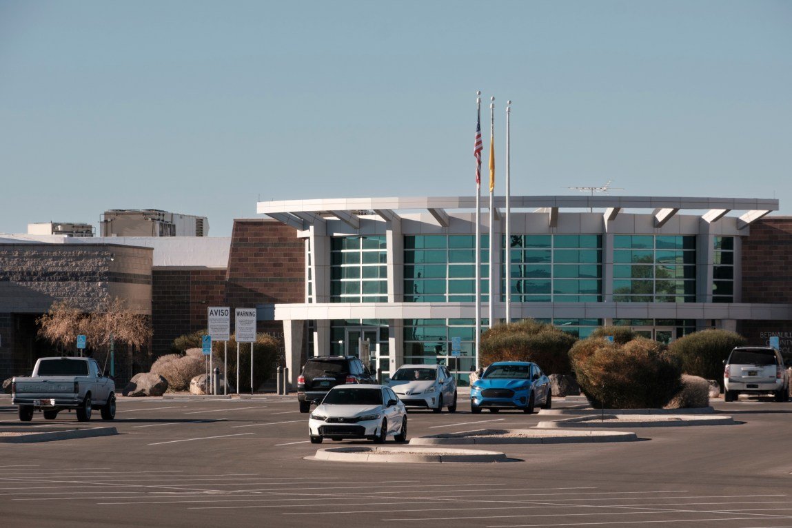 The exterior of a large building, showing an entrance, flagpoles and cars in the parking lot.