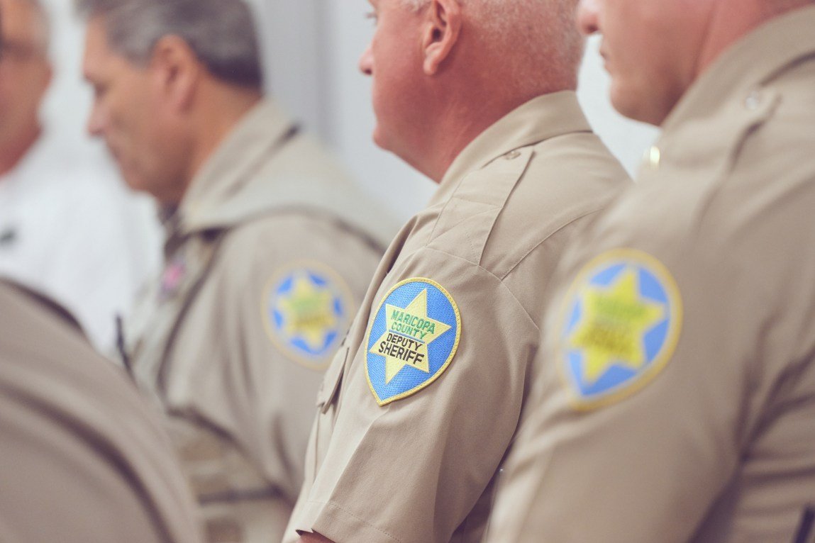 Several officers are seen from the side standing in line and wearing tan uniform shirts with prominent shoulder patches that read “Maricopa County Deputy Sheriff” and feature a yellow star on a blue background.