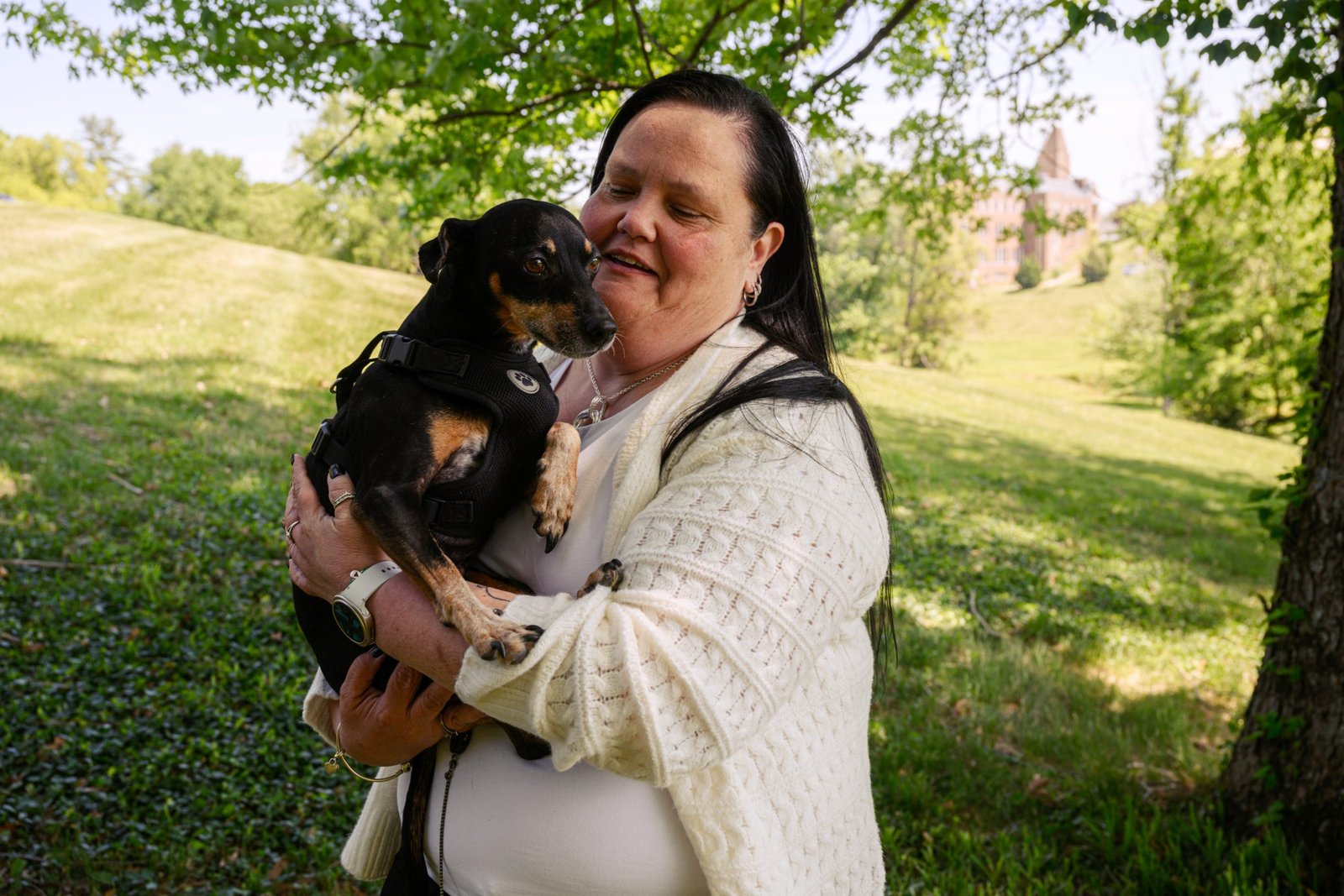 A woman with long black hair and a cream-colored knit cardigan holds a small, black-and-tan dog wearing a harness. They are outdoors under a tree in a grassy park.