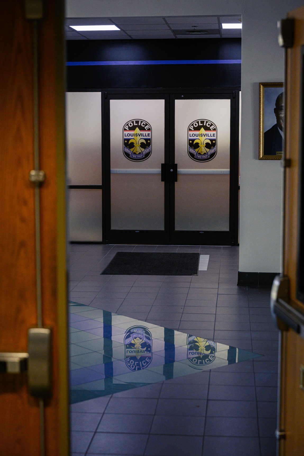 A set of frosted glass double doors features the Louisville Metro Police department badge logo on each pane, with the reflection of the logos mirrored on the polished tile floor.