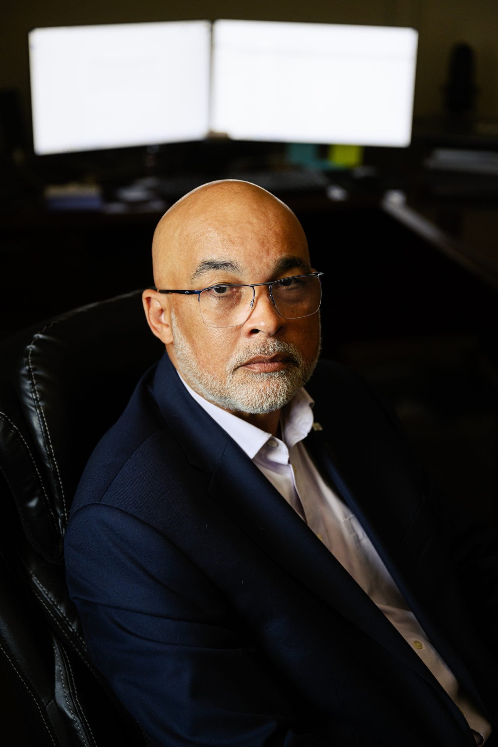 A portrait of a bald man with a gray beard and glasses, wearing a navy blue blazer and a white button-down shirt. He is sitting in a black leather office chair with a serious expression, and two illuminated computer monitors behind him.