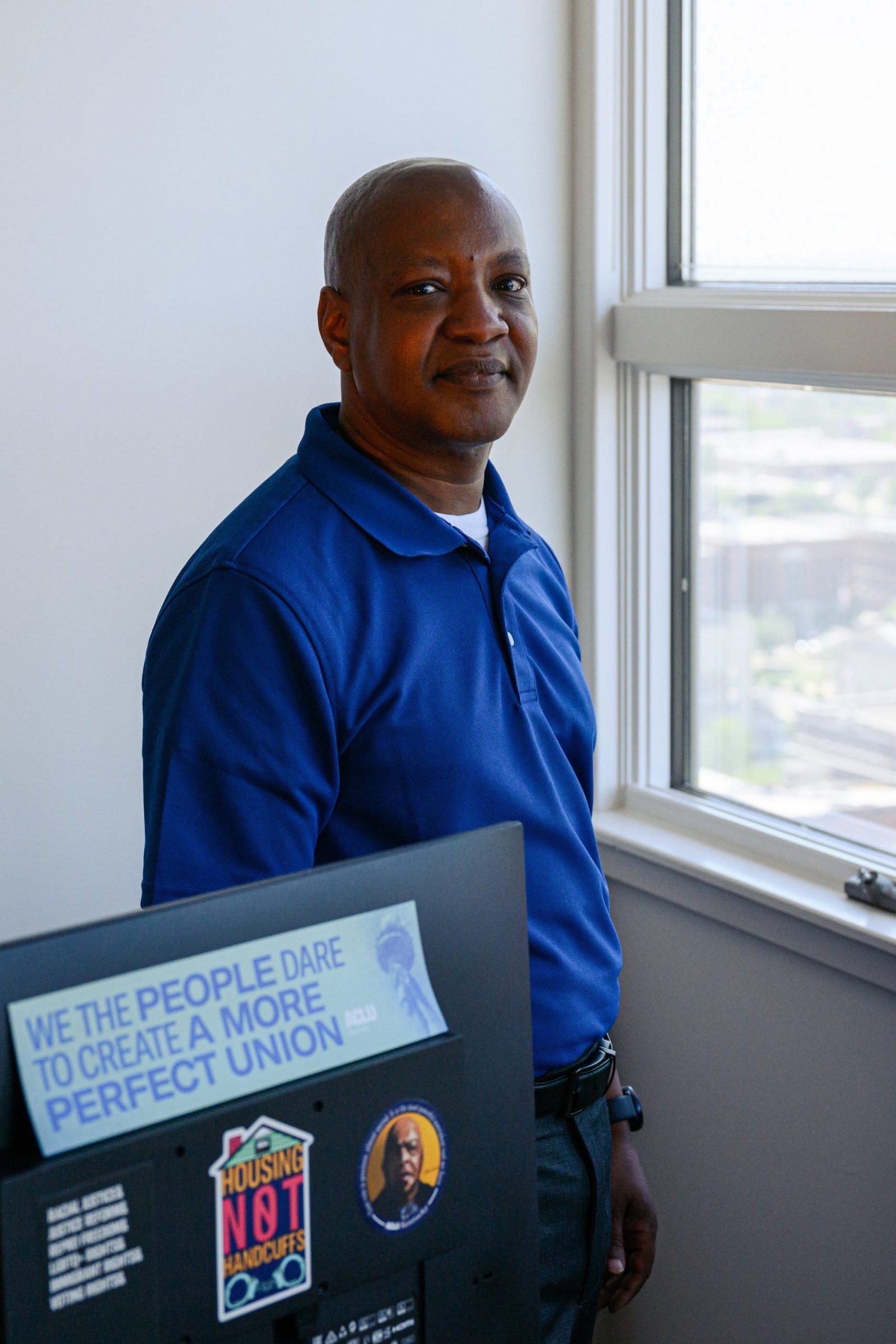 A man standing next to an office window, looking toward the camera with a gentle expression. He is wearing a royal blue polo shirt. In the foreground, the back of a computer monitor features stickers that say, “We the people dare to create a more perfect union,” and “Housing, not handcuffs.”