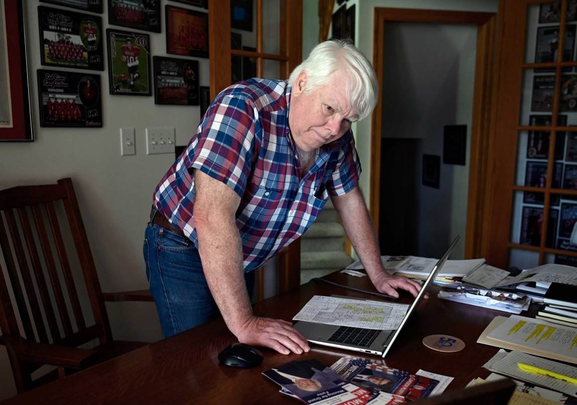 A man in a plaid shirt and jeans leans over a wooden desk, looking intently at a laptop screen surrounded by papers, maps and campaign flyers.