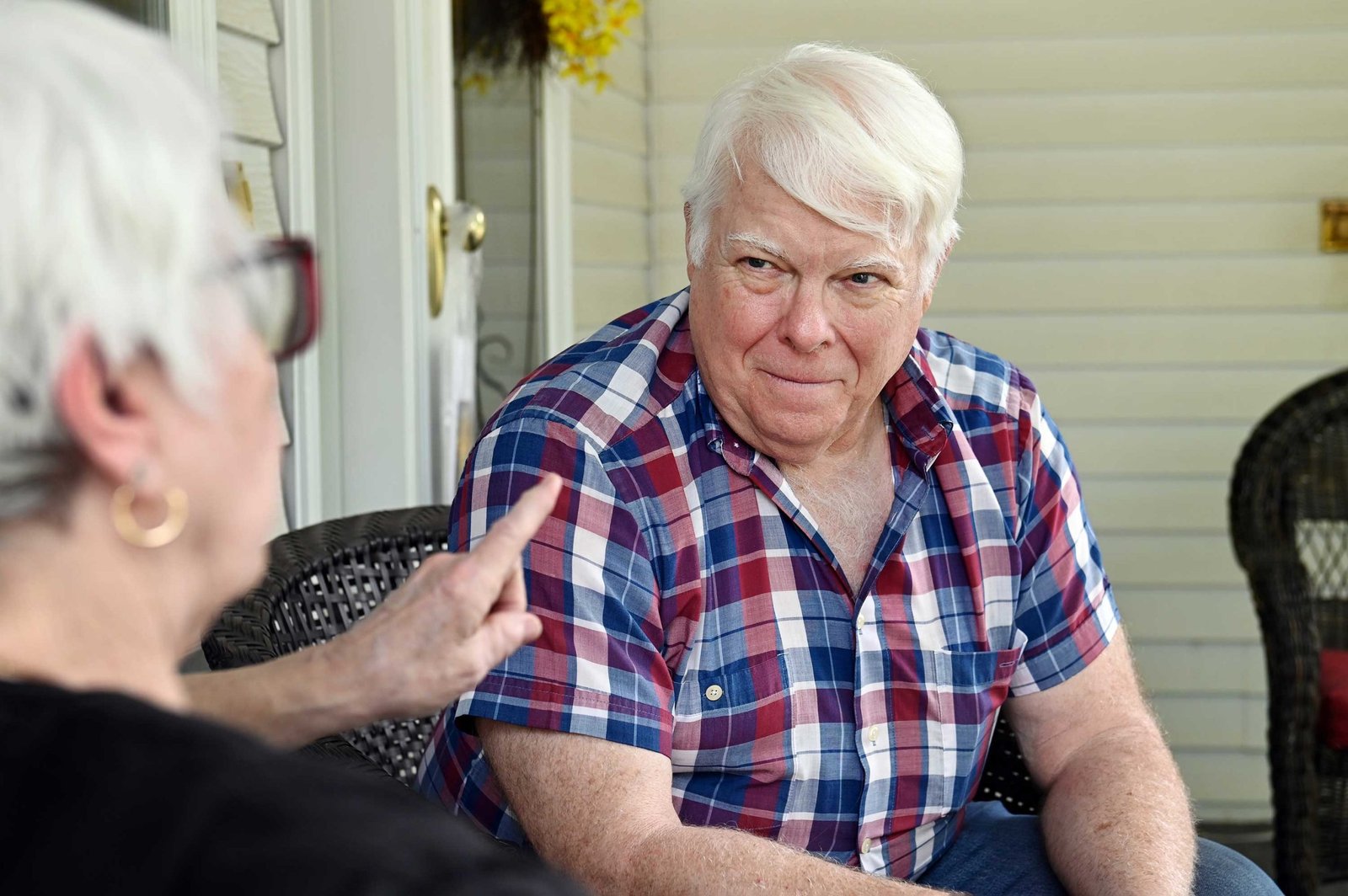 A white-haired man in a plaid shirt sits on a porch, listening intently to a woman speaking to him in the foreground.