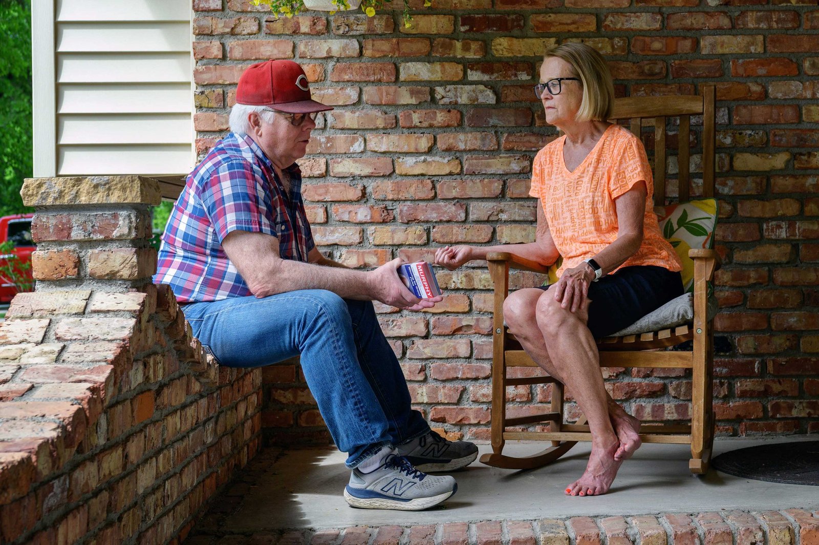 A man in a red baseball cap and plaid shirt sits on a low brick wall, passing campaign literature to a barefoot woman sitting in a rocking chair on a brick porch.