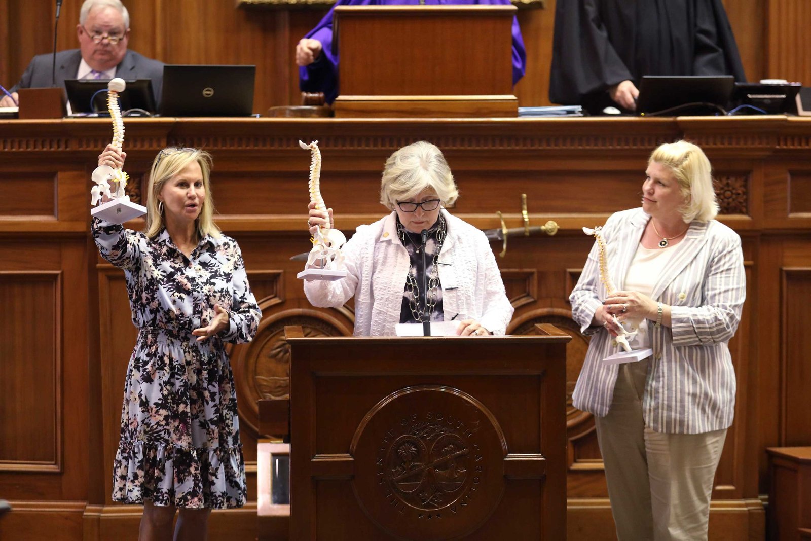 Three women stand at a legislative podium holding up anatomical models of human spines.
