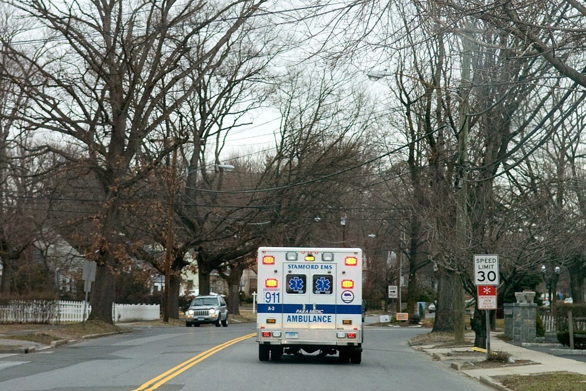 An ambulance drives down a tree-lined street. The words “Stamford EMS,” “Ambulance” and “911” are visible on the back of the vehicle.