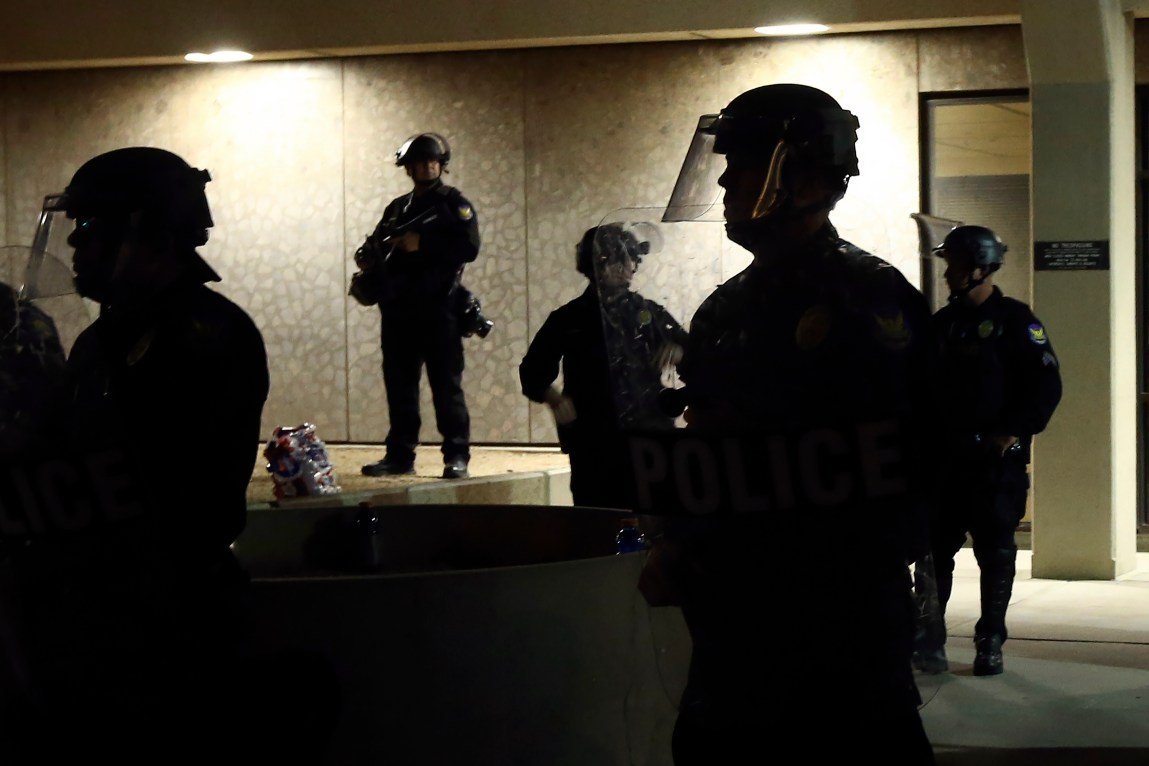 Five police officers wearing helmets with shields stand outside of a building with overhead lighting. The officers are silhouetted and anonymous.