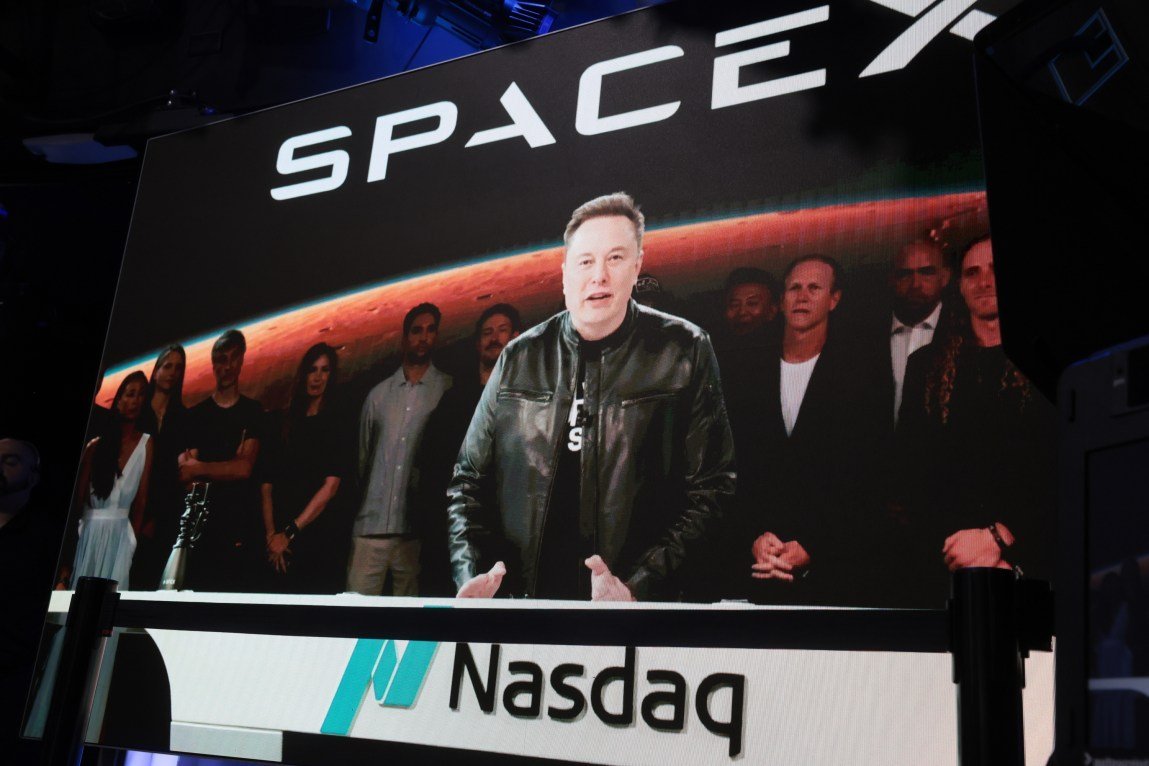 Elon Musk speaking on a screen with a crowd of people behind him. Above him is the SpaceX logo and below him is the Nasdaq logo.