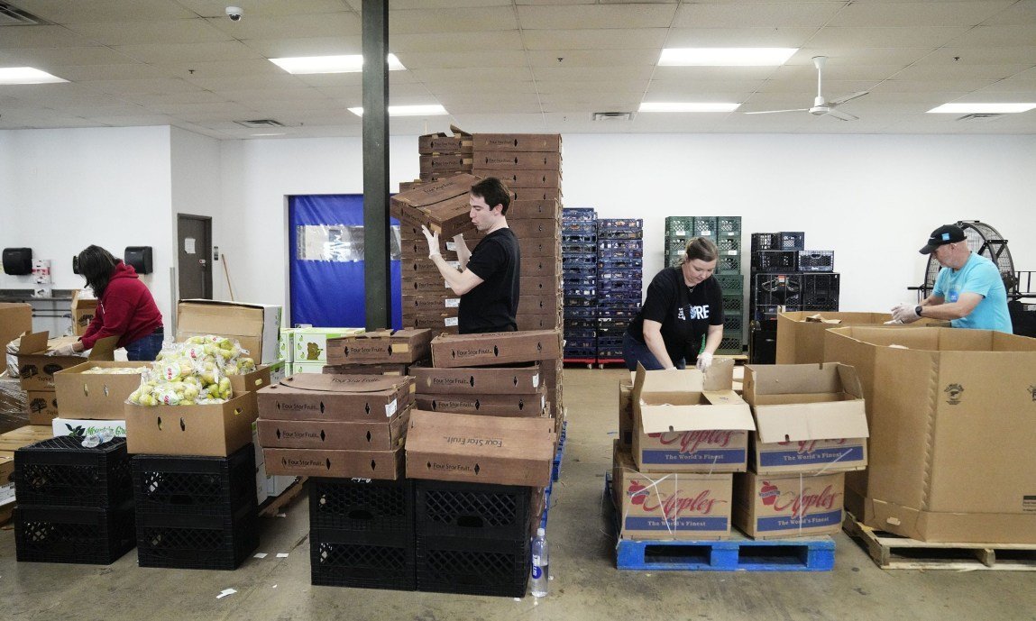 Volunteers in a large warehouse space organizing, stacking and packing cardboard produce boxes labeled with various brand names.