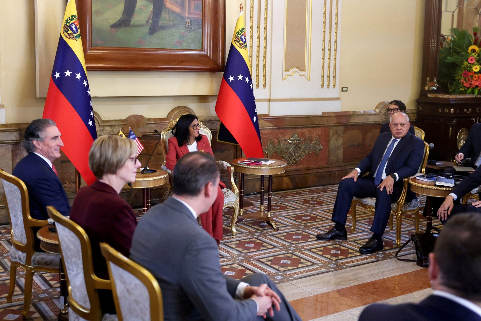 Several seated men and women in suits smile at one another during a meeting, in a room in a government building with two Venezuelan flags.