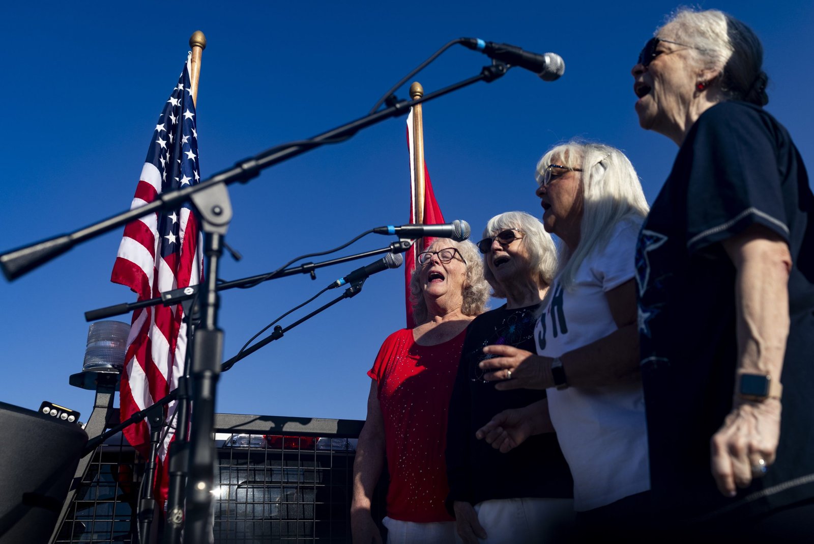 Four women stand side by side singing into microphones outdoors during a daytime event. Flags flank them in the background against a clear, deep blue sky.