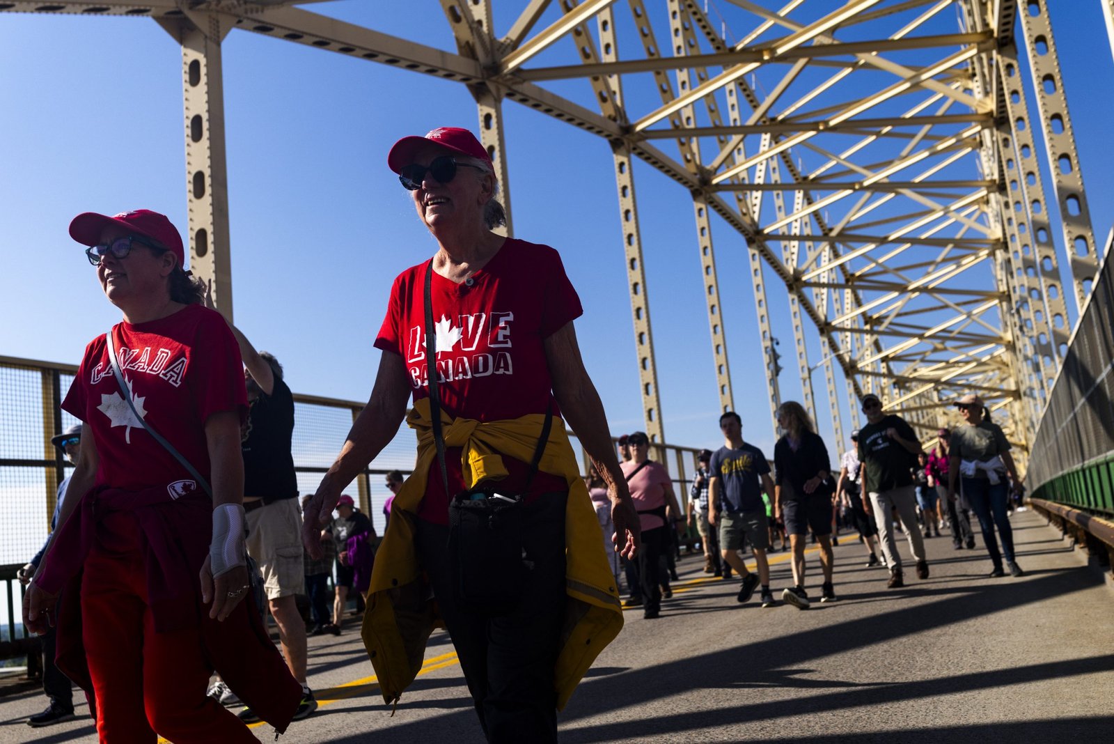 Two women wearing matching red Canada-themed shirts and red caps walk across a bridge amid a crowd of pedestrians. The sunlit bridge frame towers overhead against a clear blue sky.