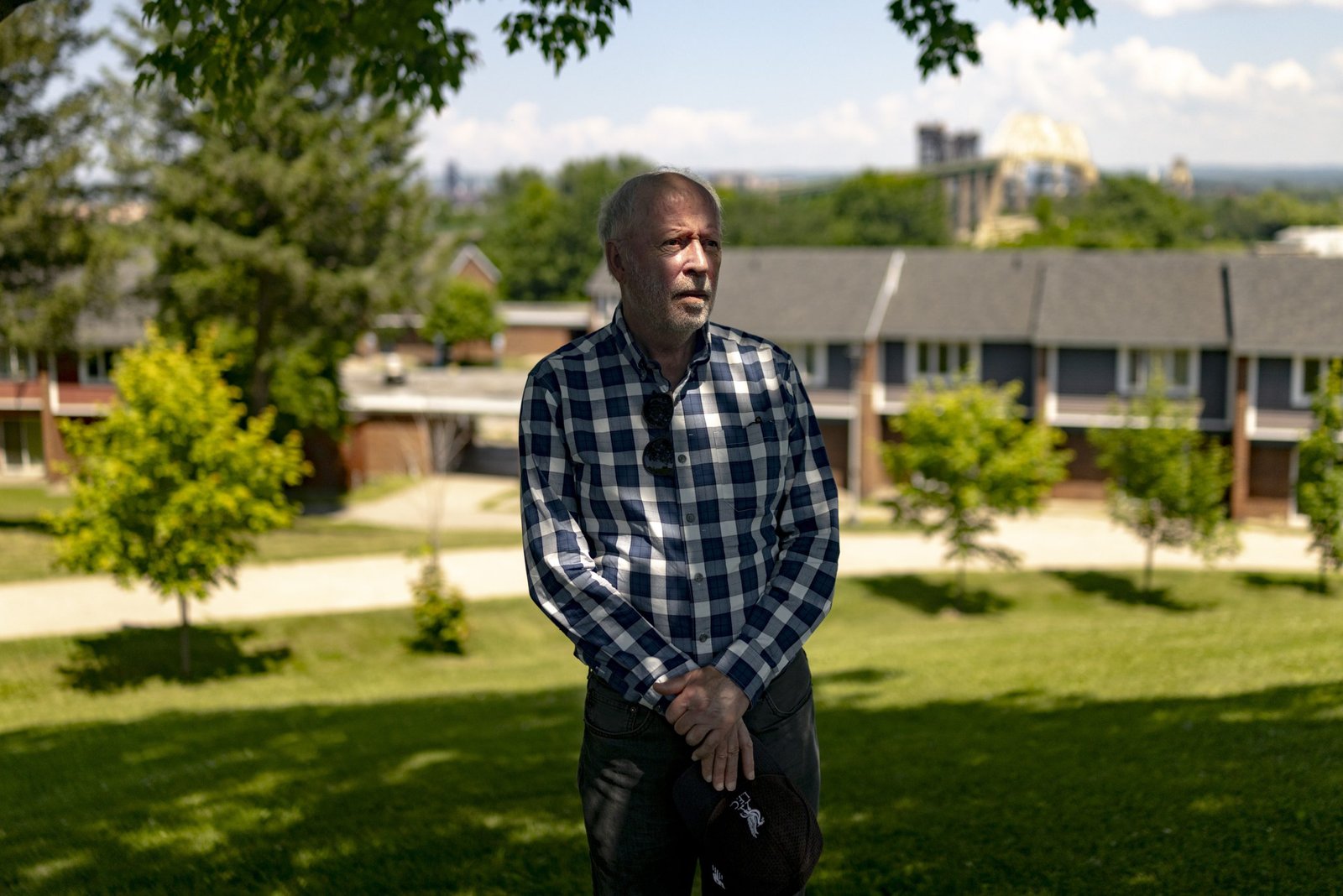 An older man with graying hair and a beard stands outdoors in a grassy area, wearing a blue-and-white plaid button-up shirt and dark pants. He rests his hands together in front of him, holding a dark cap.
