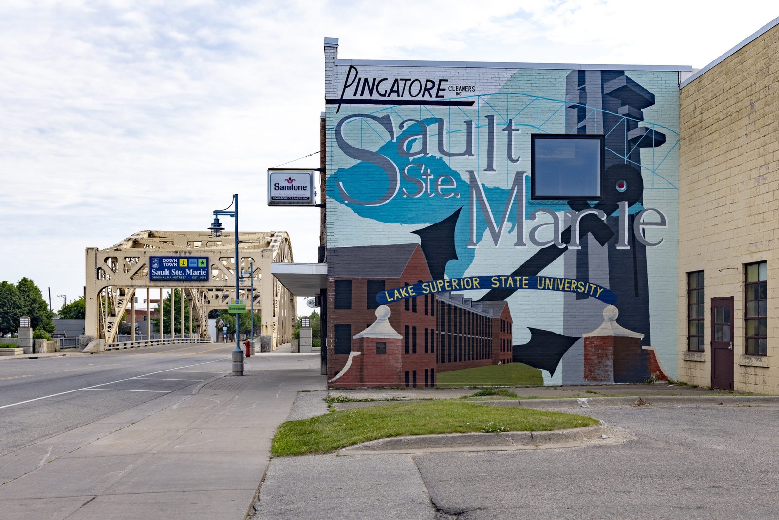 A street-level view shows a building adorned with a large Sault Ste. Marie mural that includes the phrases “Pingatore Cleaners Inc.” and “Lake Superior State University.” In the background, a light-yellow steel bridge spans a road under a partly cloudy sky.