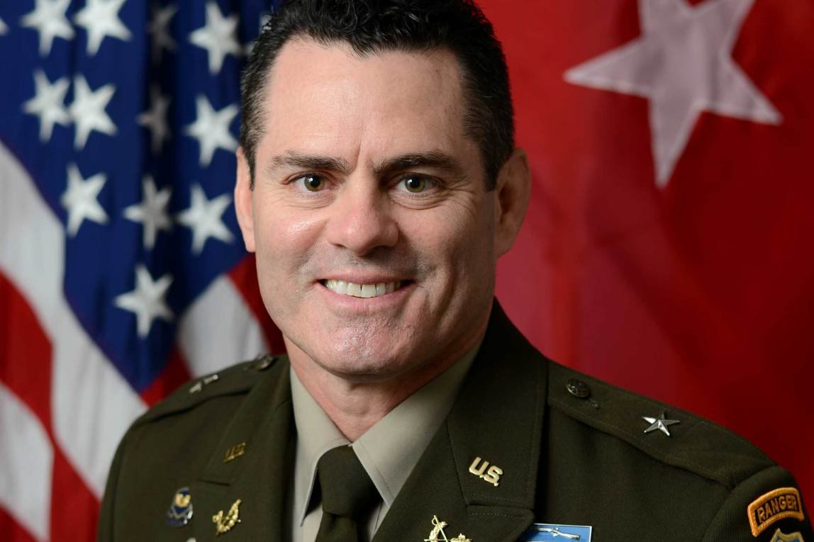 An official military portrait shows a man with dark, short hair smiling directly at the camera. He is wearing a dark olive-green U.S. Army uniform with a light-colored dress shirt and dark tie.