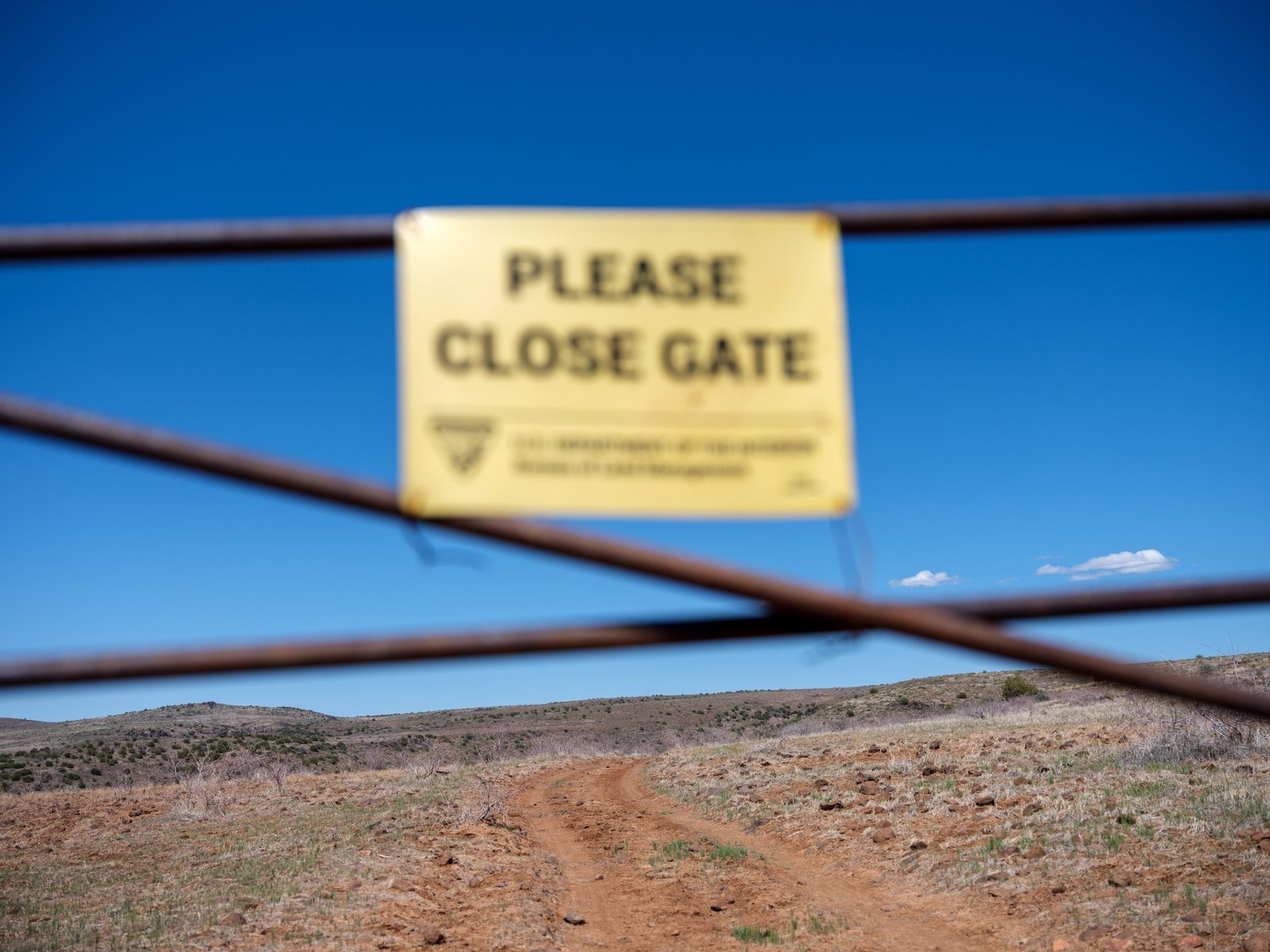 A blurred yellow sign reading “Please close gate” is mounted on a wire fence in the foreground. Through the fence, a sharp view reveals a winding dirt road cutting through a dry, hilly landscape under a vivid blue sky.