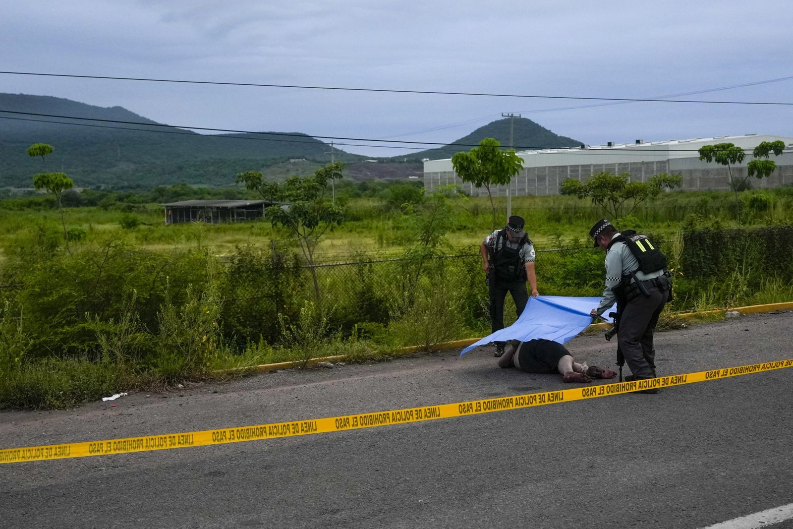Two people in uniform drape a tarp over a dead body behind yellow police tape on the side of a road. The background is a lush green field with mountains in the distance.