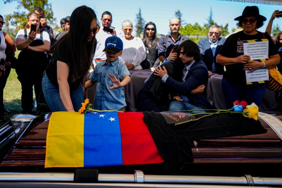 A funeral scene in which a coffin is draped with a Venezuelan flag. Above it, a woman and a boy reach their hands toward the coffin. In the background, two people embrace, and a crowd of people looks on.