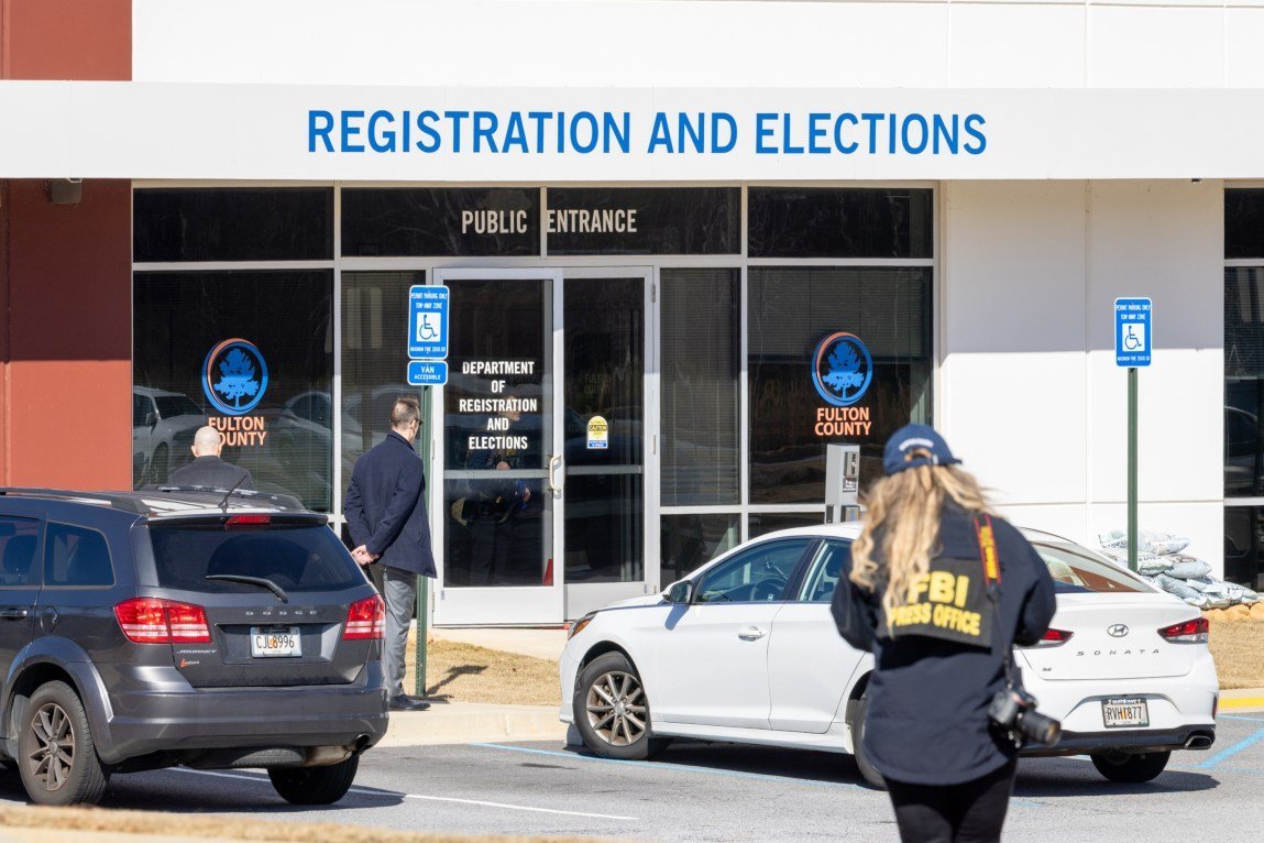 An FBI personnel member with “FBI press office” written on their jacket walks toward the public entrance of the Fulton County Department of Registration and Elections building. Two parked cars sit in the foreground, and two individuals stand near the glass doors of the entrance under a sign reading “registration and elections.”
