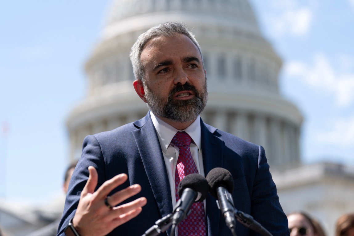 A man in a suit and tie gestures with his hand as he speaks into two microphones. The U.S. Capitol is in the background.