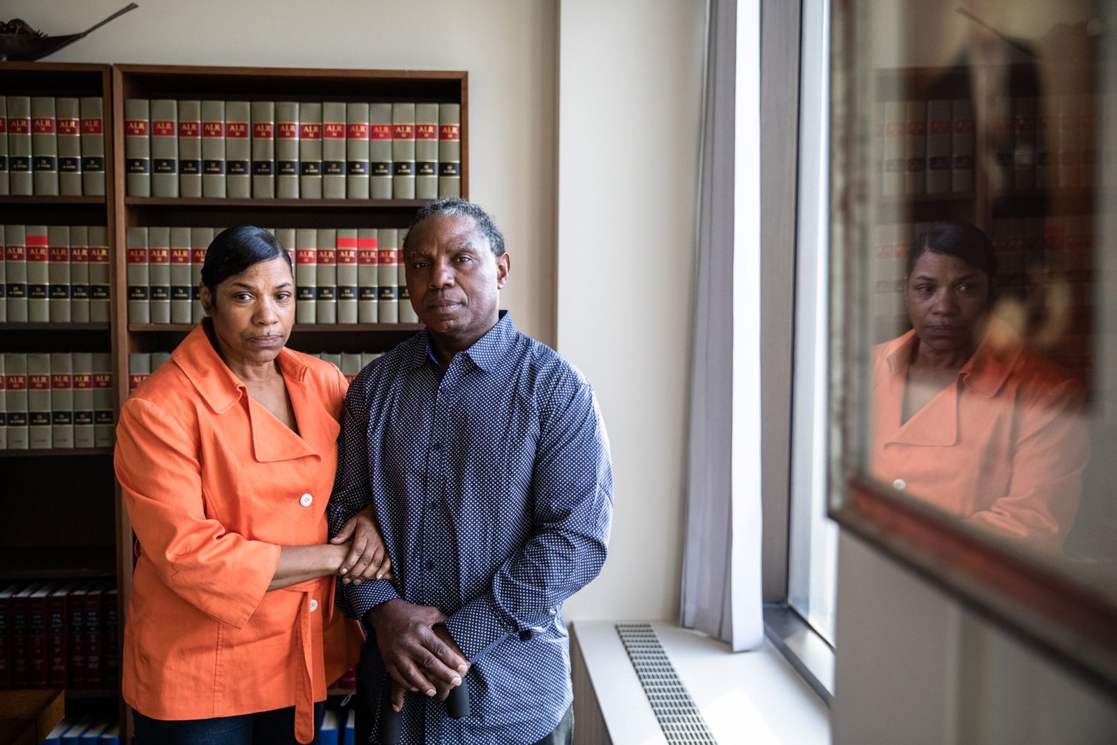 A man and a woman stand together with somber expressions, looking toward the camera. The woman is wearing a bright orange jacket and holds the arm of the man, who is wearing a blue patterned shirt. A bookshelf filled with legal volumes stands behind them, and a glass-framed reflection is visible on the right.