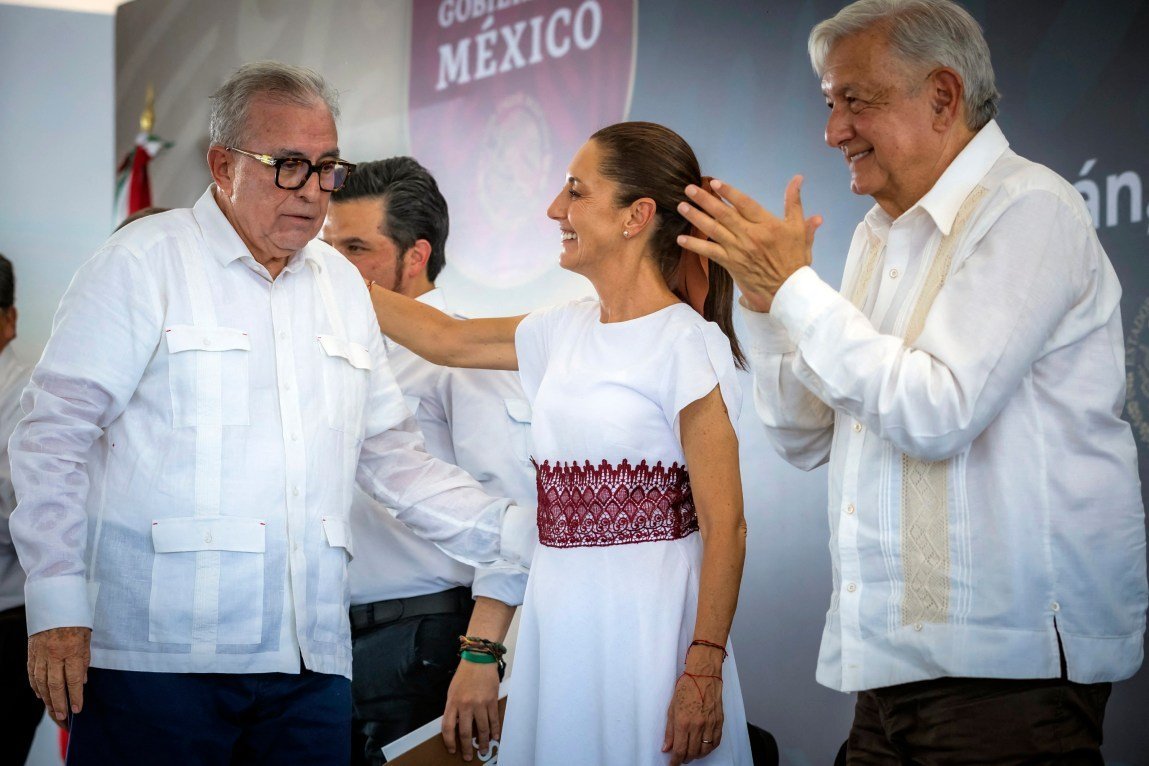 A man and a woman reach out to embrace each other as another man applauds at a government event.
