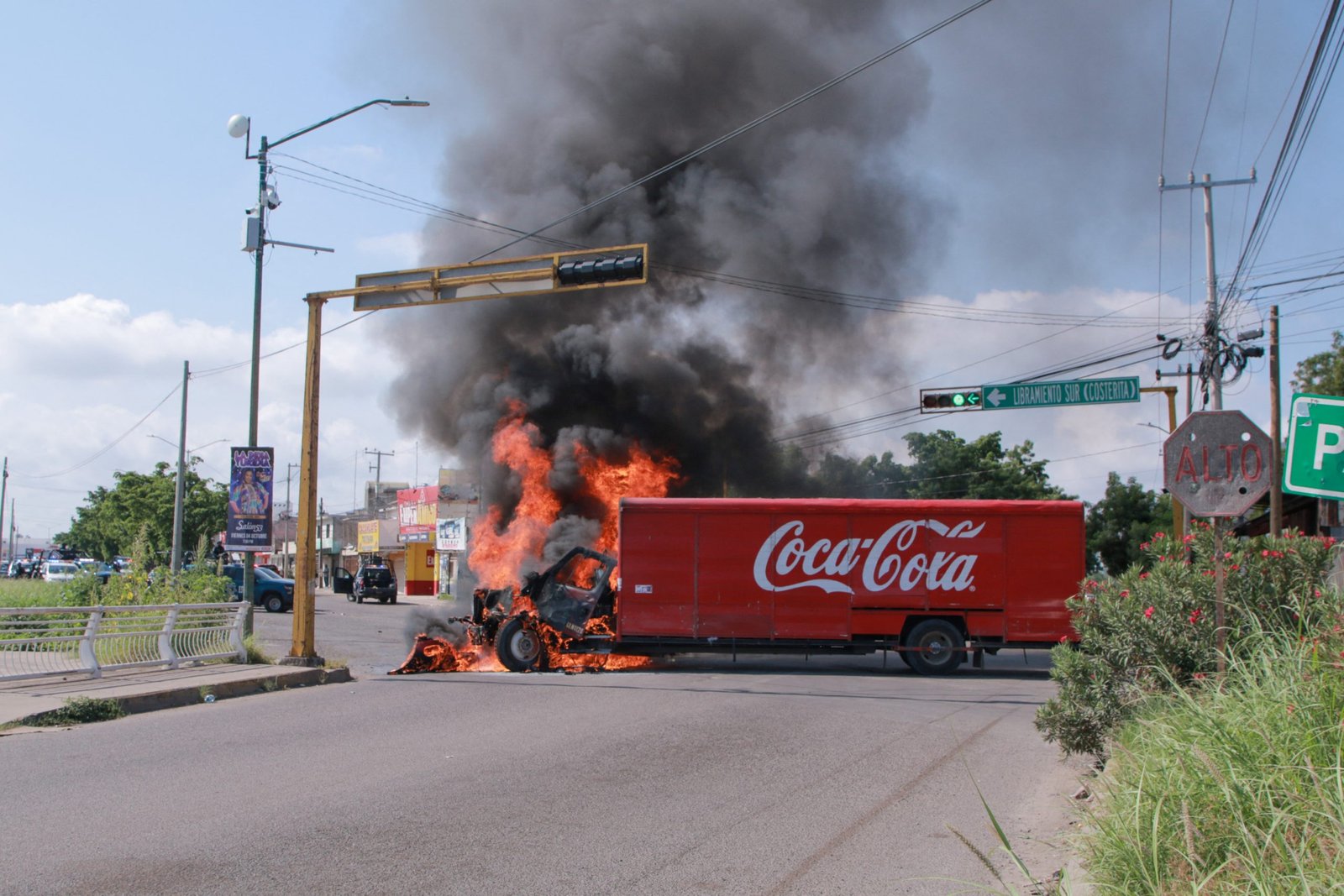 The cab of a large truck is on fire, with flames going high in the air and black smoke in the sky. The truck is red, and “Coca-Cola” is written on the side. It is in the middle of an intersection in a city street.