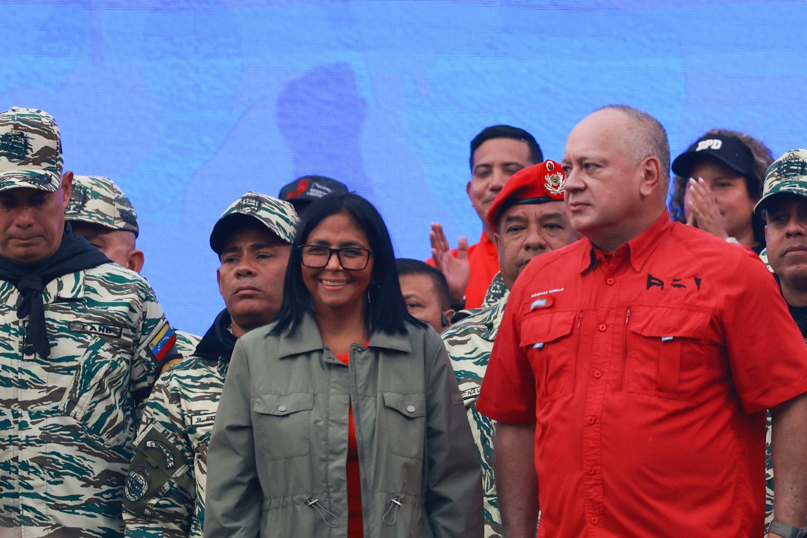 A smiling woman and a man with a serious expression stand in front of a group of people, most of whom are wearing military uniforms.