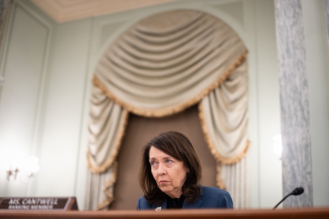 A woman with shoulder-length brown hair sits behind a wooden table with a microphone and a placard reading “Ms. Cantwell.” The room has an ornate curtain hanging behind her and high white ceilings.