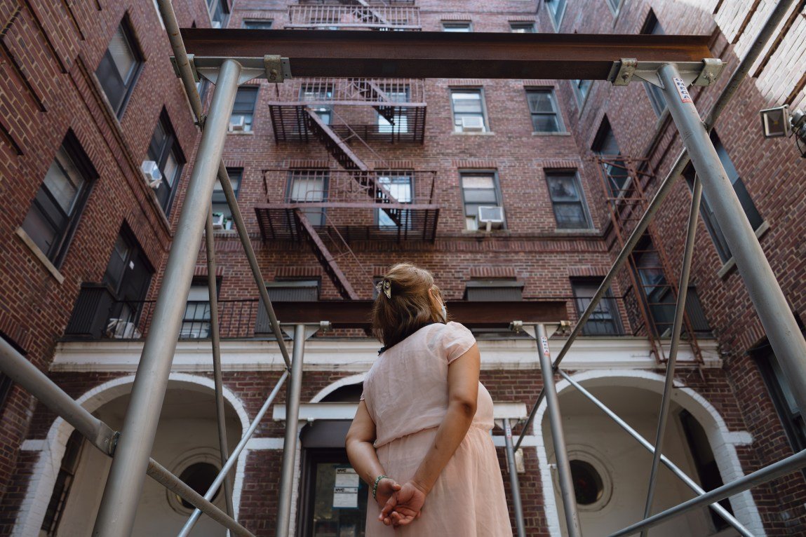 A woman stands outside a brick building, looking away from the camera, with her hands clasped behind her back.