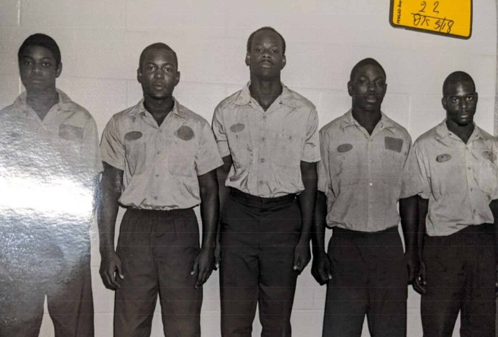 A black-and-white police lineup photograph shows five men standing side by side against a cinder block wall. They are wearing matching short-sleeved shirts and trousers, looking directly at the camera.