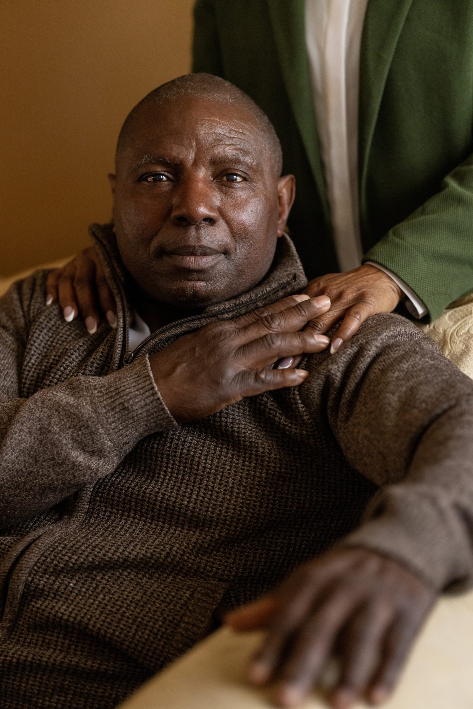 A man in a brown sweater sits on a chair looking at the camera. A person behind him rests their hands on his shoulders, and he has his hand placed on top of theirs.