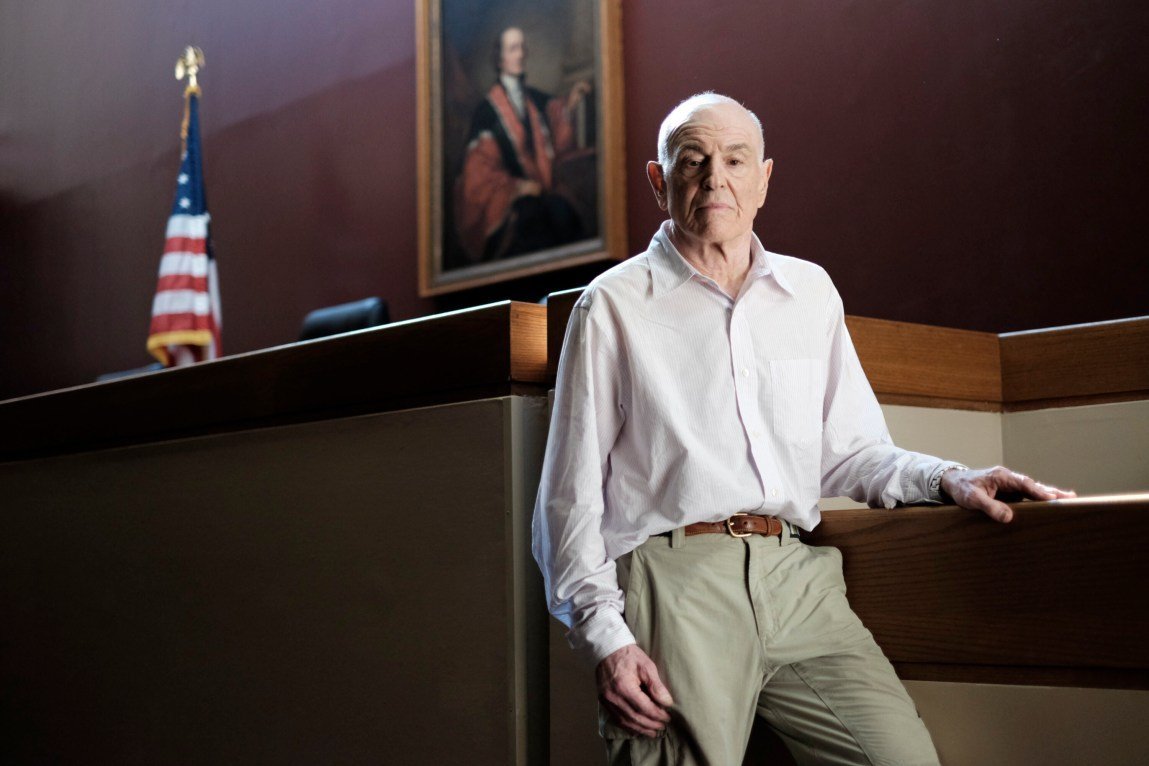 A man with white hair who is wearing a collared shirt rests his arm on the witness stand in a courtroom.