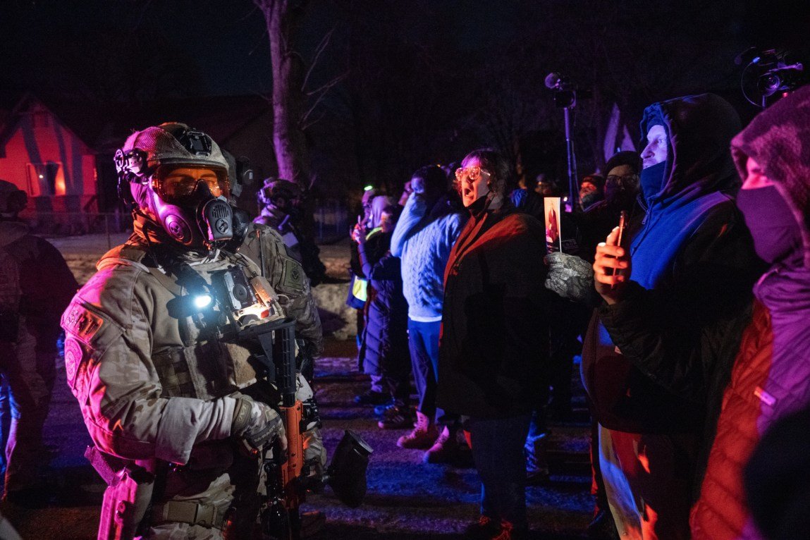 A line of protesters in winter clothing film and shout at law enforcement officers who are wearing tactical gear and holding paintball guns.